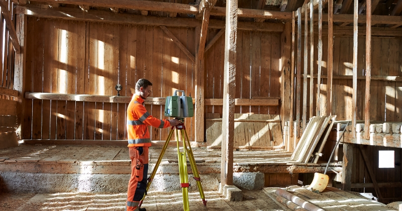 Mitarbeiter der Gossweiler Ingenieure vermisst eine alte Holz-Scheune mit einem Laserscanning-Gerät, das auf einem Stativ montiert ist.