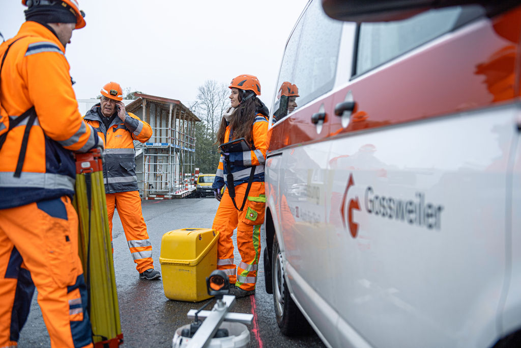 Mitarbeiterin der Gossweiler Ingenieure in oranger Bekleidung und Helm lehnt sich an das Firmenfahrzeug und unterhält sich mit dem Bauleiter.