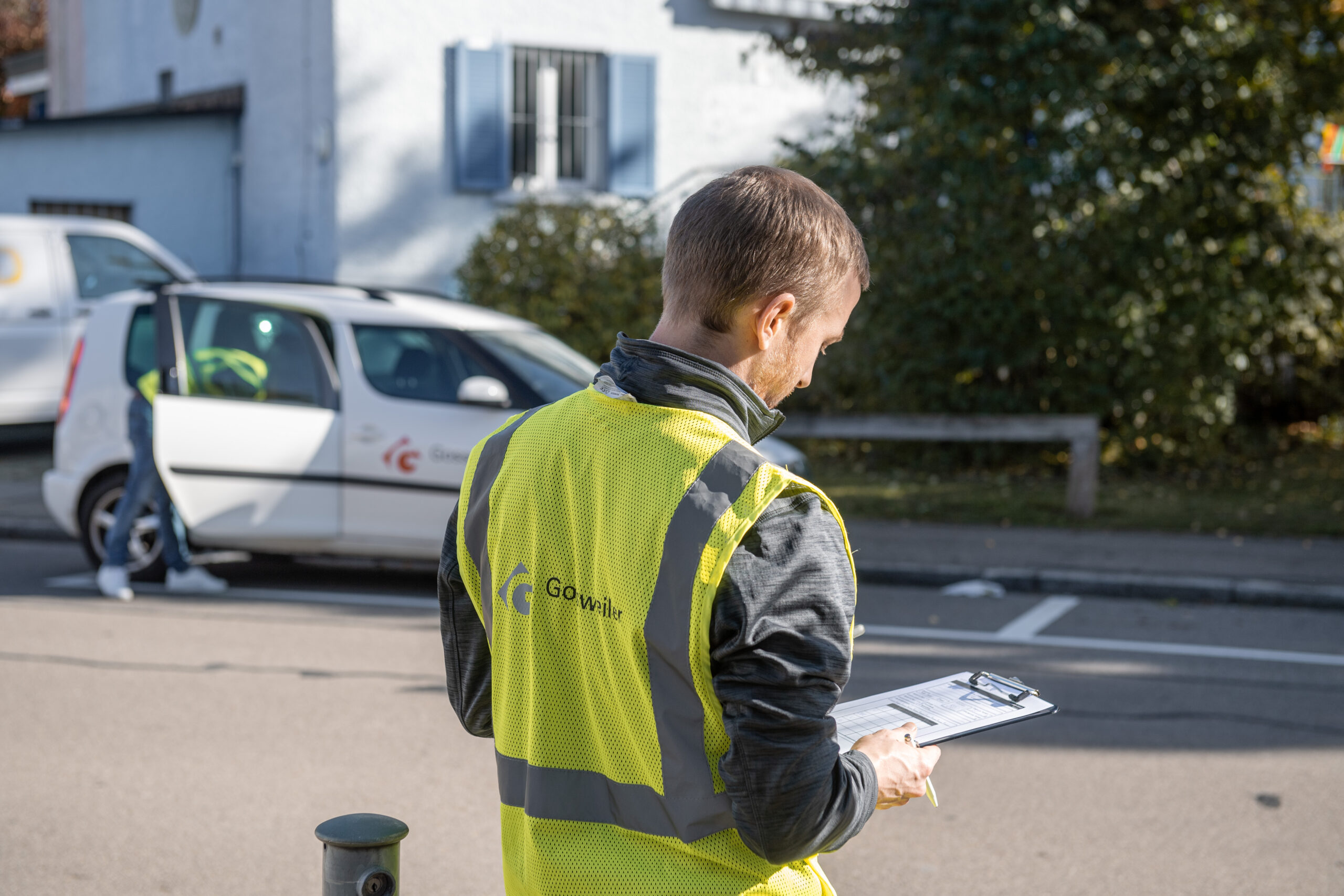 Mitarbeiter der Gossweiler Ingenieure in gelber Weste steht am Strassenrand, schaut auf seine Daten. Im Hintergrund lädt ein Mitarbeiter etwas aus dem Firmenfahrzeug.